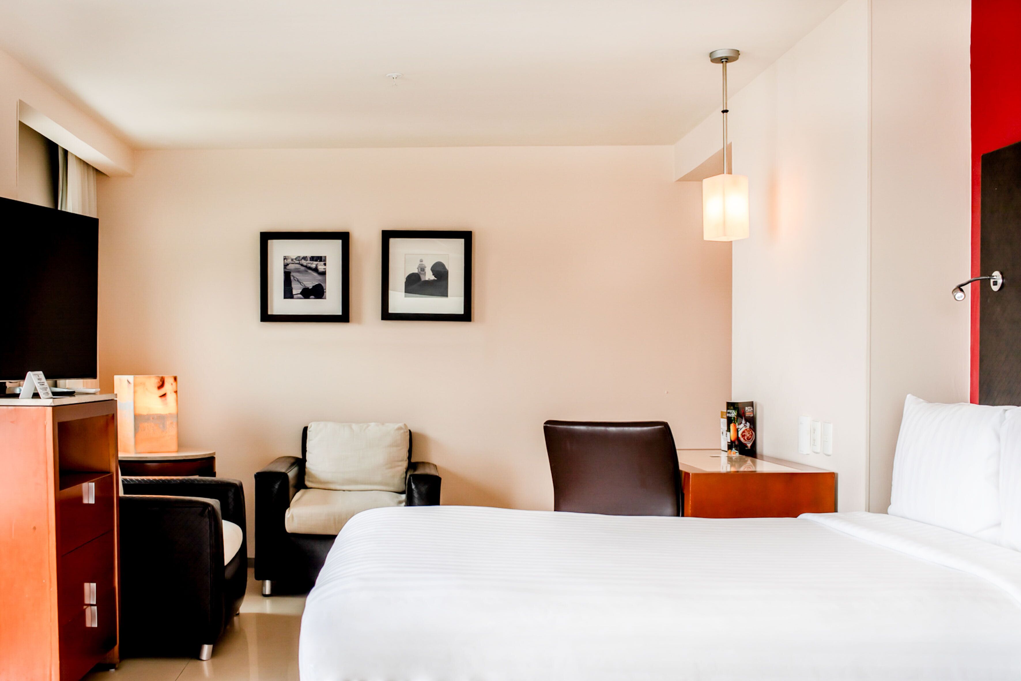 A hotel room with a bed, two chairs, a desk, a TV, and two framed pictures on the wall.