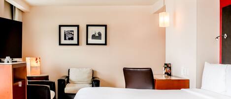 A hotel room with a bed, two chairs, a desk, a TV, and two framed pictures on the wall.