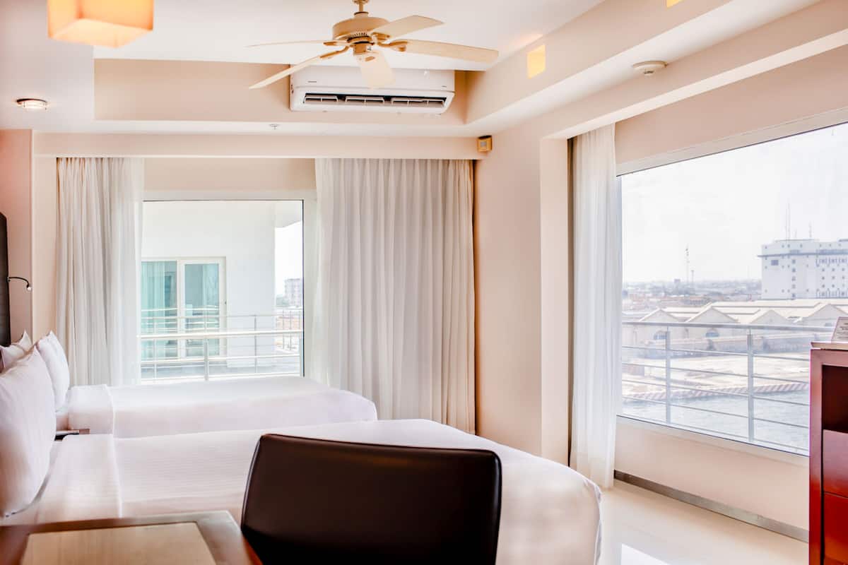 Habitación de hotel con dos camas, ventilador de techo, aire acondicionado y un amplio ventanal con vistas a la ciudad.