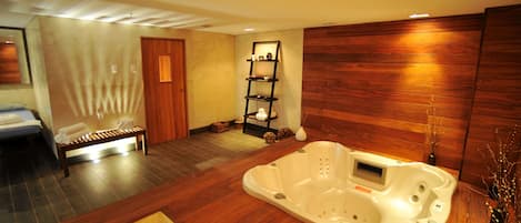 A spa room with a wooden floor, a massage table, a white bathtub, and a shelf with toiletries.