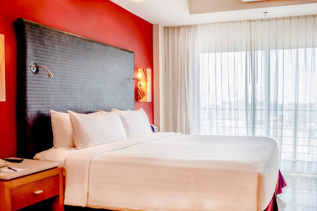 Habitación de hotel con una cama grande, una mesita de noche, una lámpara de pared y un ventanal con cortinas translúcidas.