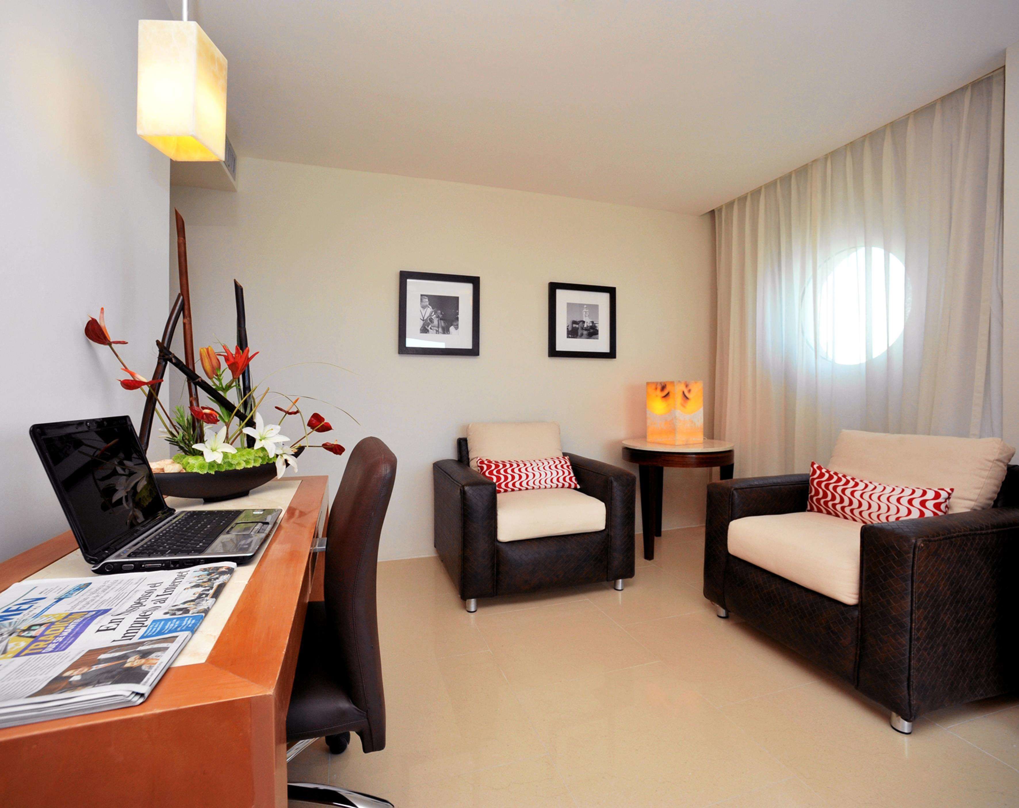A hotel room with a desk, two armchairs, a laptop, a vase with flowers, a framed picture, and a lamp.