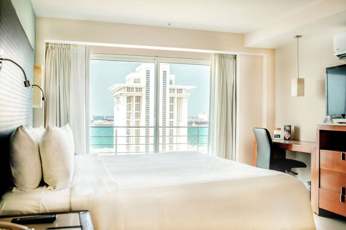 Habitación de hotel con una cama grande, un escritorio con computadora, un televisor y vista a un edificio al otro lado del agua.