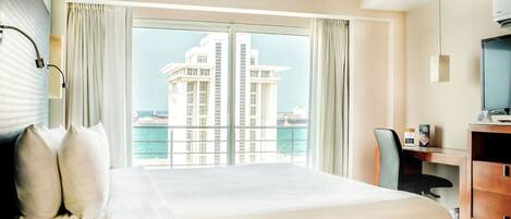 Habitación de hotel con una cama grande, un escritorio con computadora, un televisor y vista a un edificio al otro lado del agua.