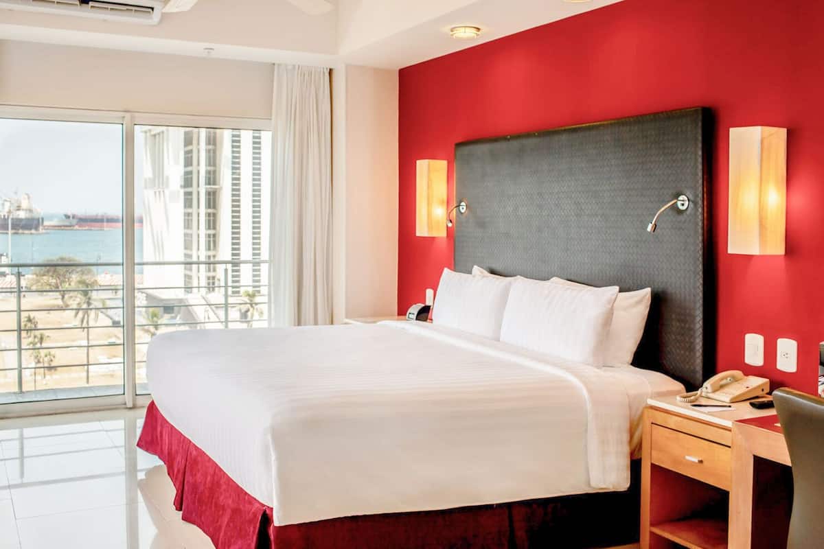 Habitación de hotel con una cama grande, un escritorio con teléfono y vistas al mar a través de amplios ventanales.