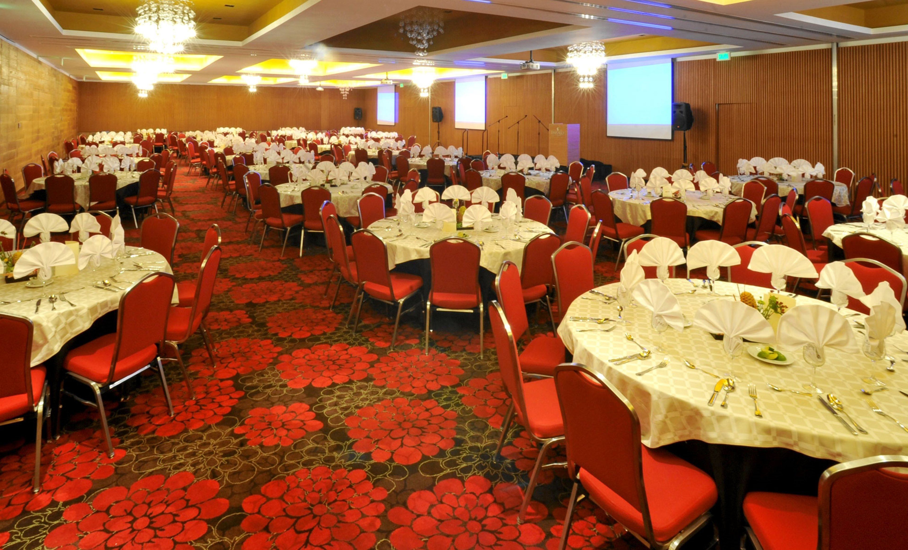 Un amplio salón de banquetes con mesas redondas dispuestas para comer, sillas rojas y una alfombra con estampado floral en rojo.