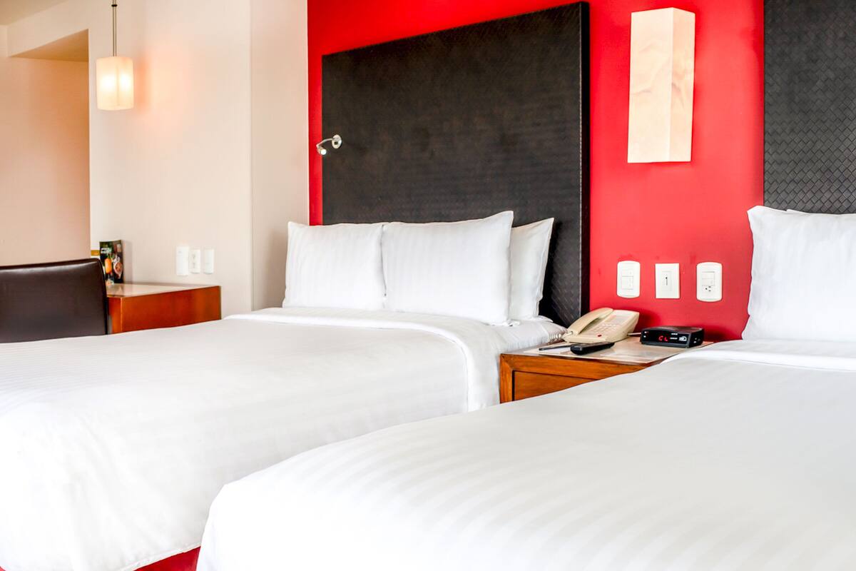 Habitación de hotel con dos camas, una pared de acento roja, una mesita de noche con teléfono y una lámpara.