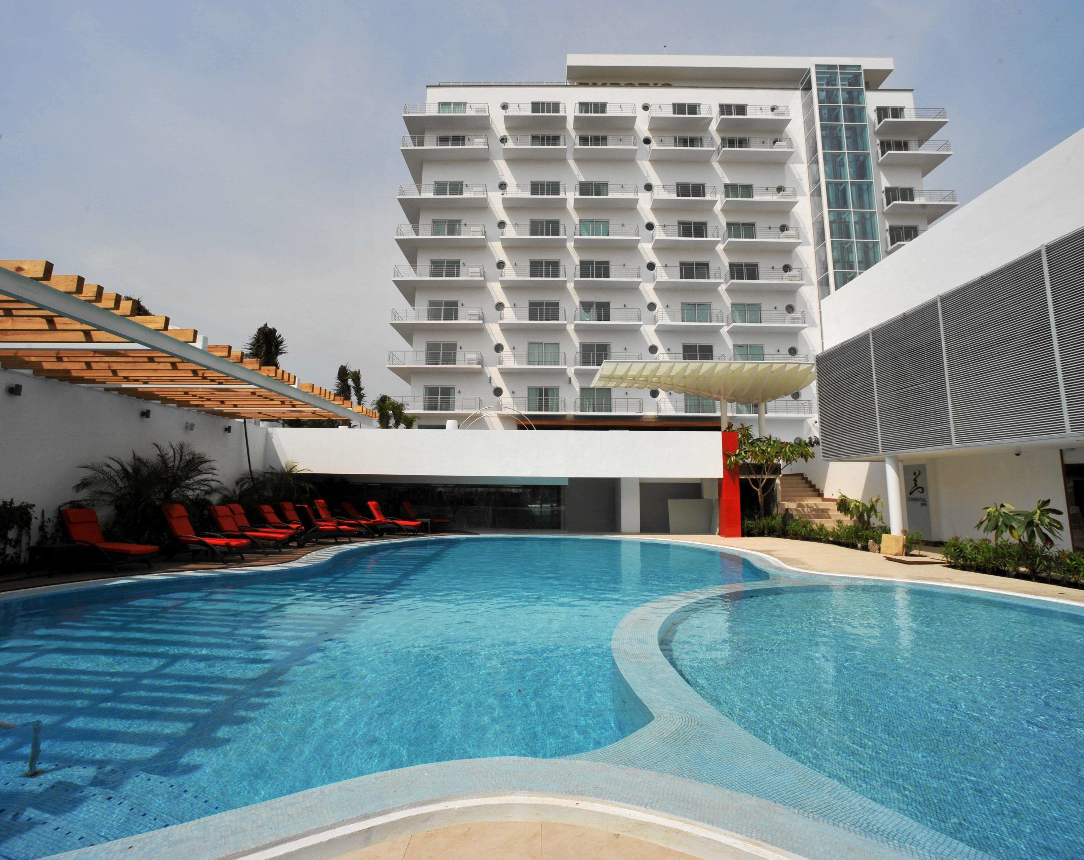 Un hotel moderno con piscina, sillas de descanso rojas y un edificio de varios pisos con balcones.