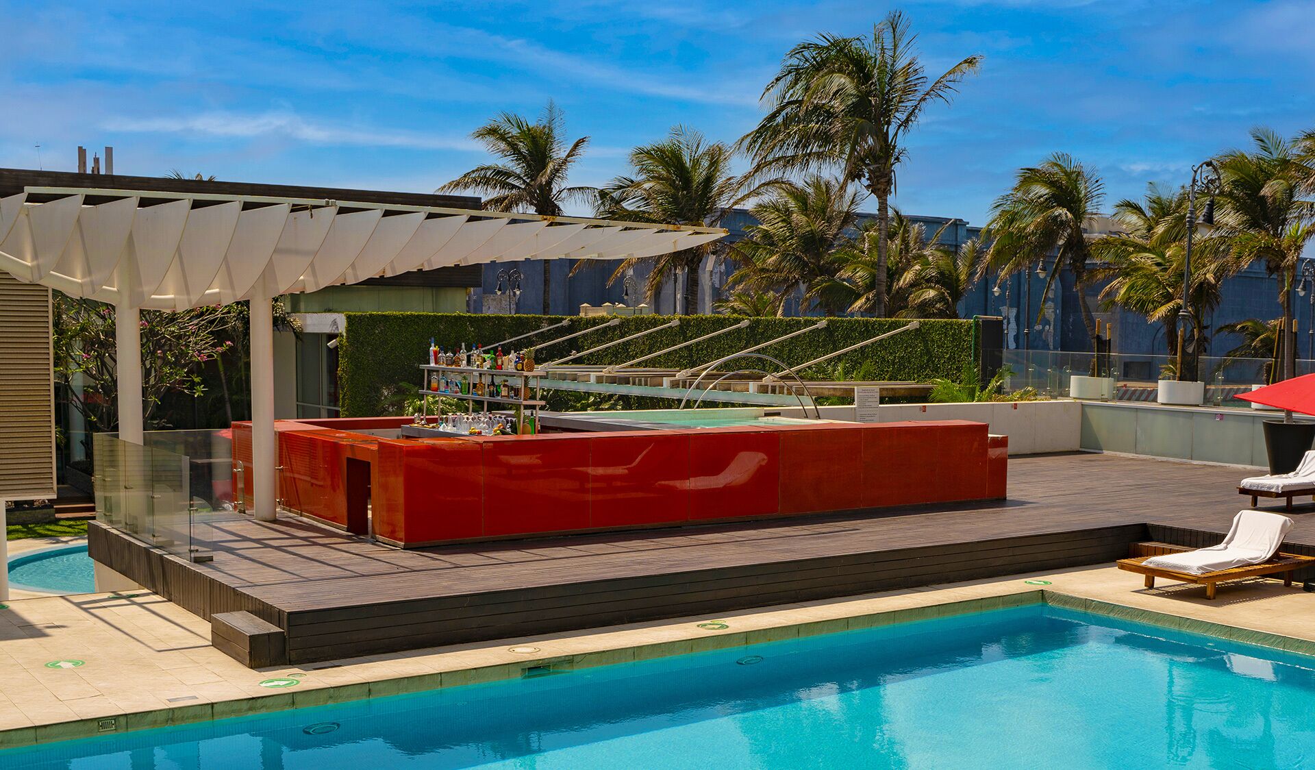 Zona de bar junto a la piscina con mostrador rojo, área de estar cubierta y una piscina con agua cristalina.