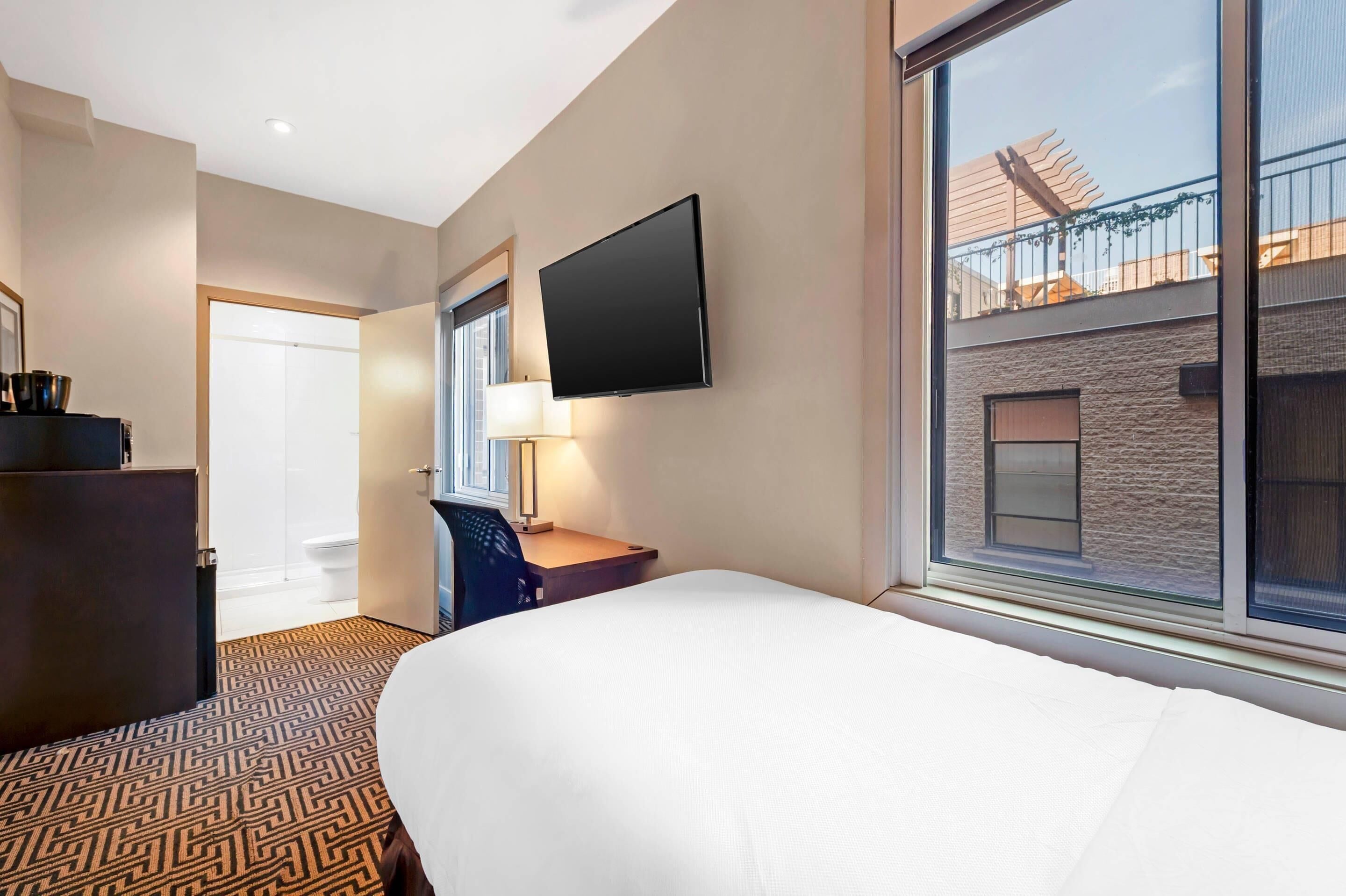 A hotel room with a bed, a desk with a chair, a TV mounted on the wall, and a window with a view of the building's exterior.
