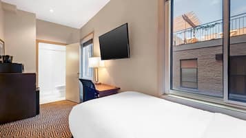 A hotel room with a bed, a desk with a chair, a TV mounted on the wall, and a window with a view of the building's exterior.