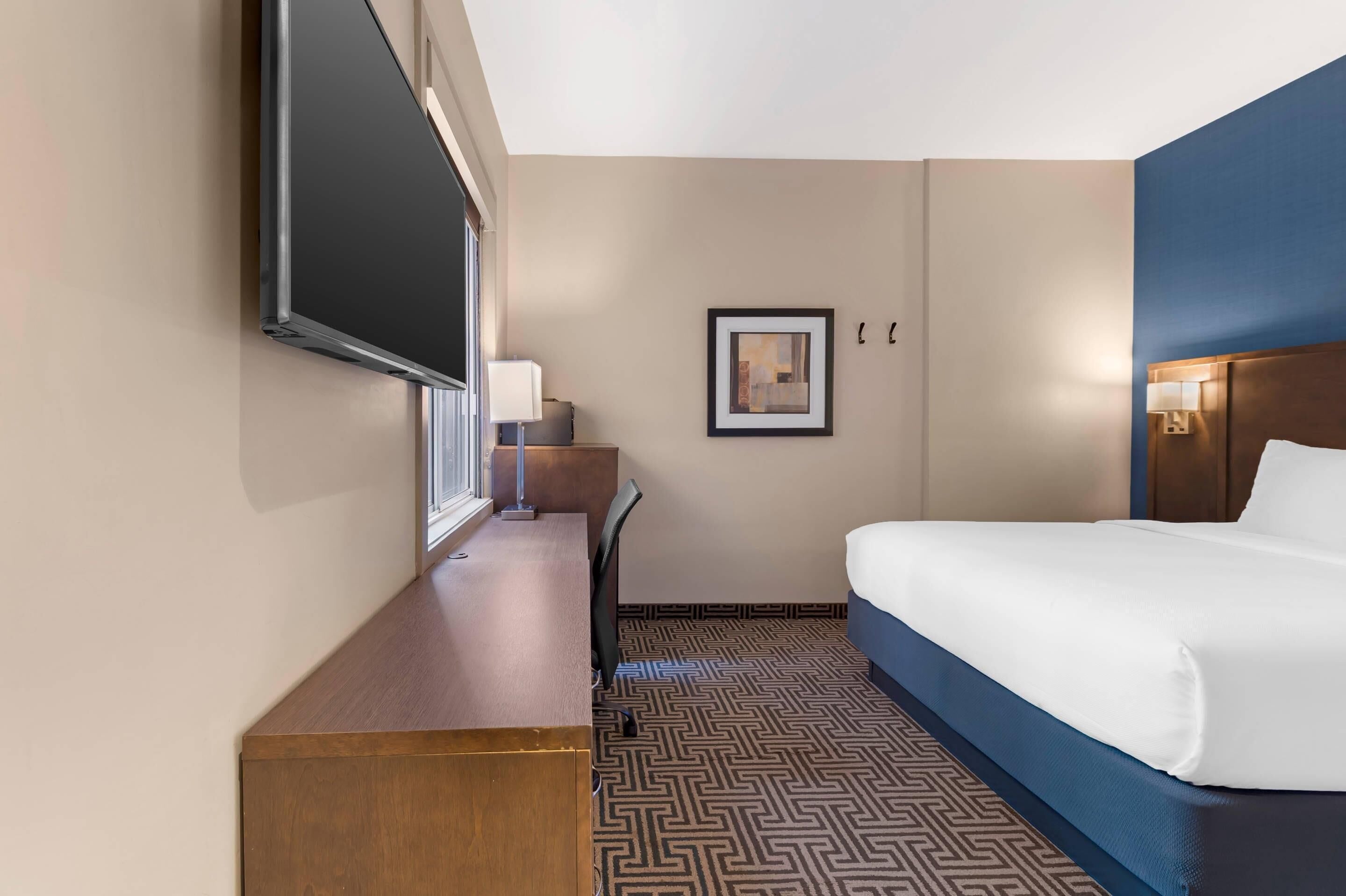 A hotel room with a bed, a desk, a chair, a TV, and a framed picture on the wall.