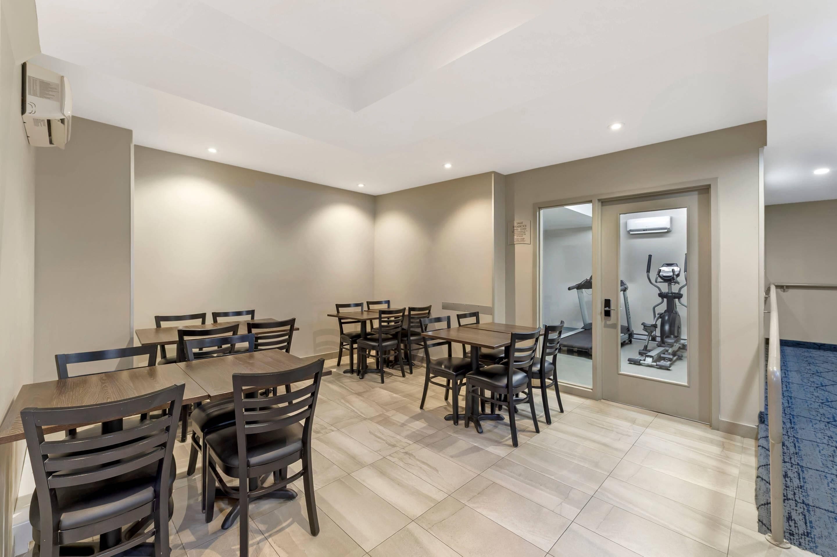 Un espace repas avec plusieurs tables et chaises, un plafond doté d’un éclairage encastré et une vue sur le matériel de musculation, visible à travers une porte vitrée.