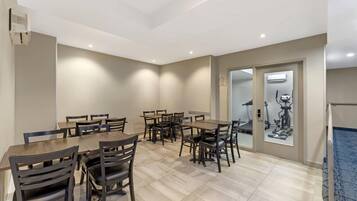 Un espace repas avec plusieurs tables et chaises, un plafond doté d’un éclairage encastré et une vue sur le matériel de musculation, visible à travers une porte vitrée.