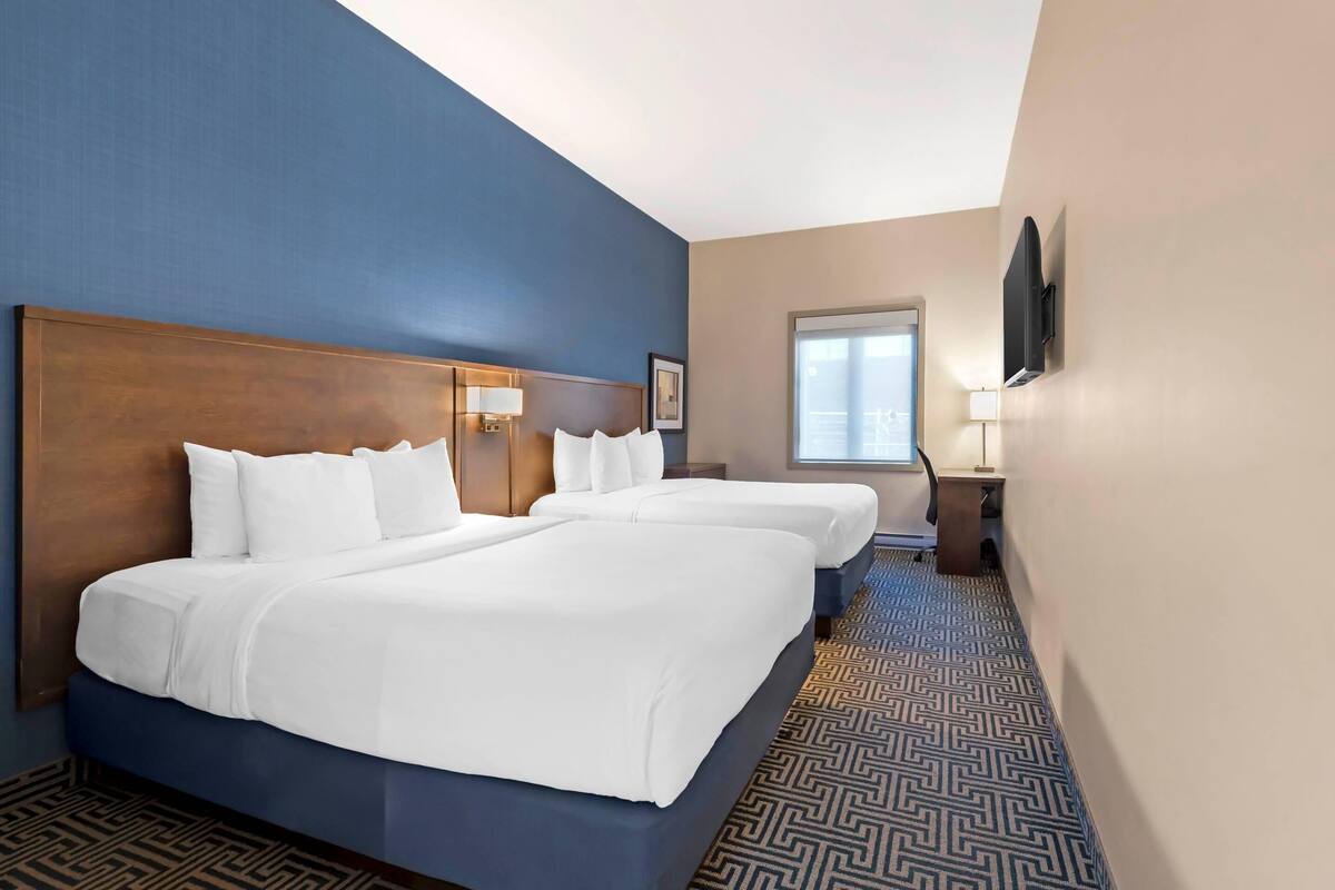 A hotel room with two beds, a large wooden headboard, a patterned carpet, a window, and a flat-screen TV mounted on the wall.
