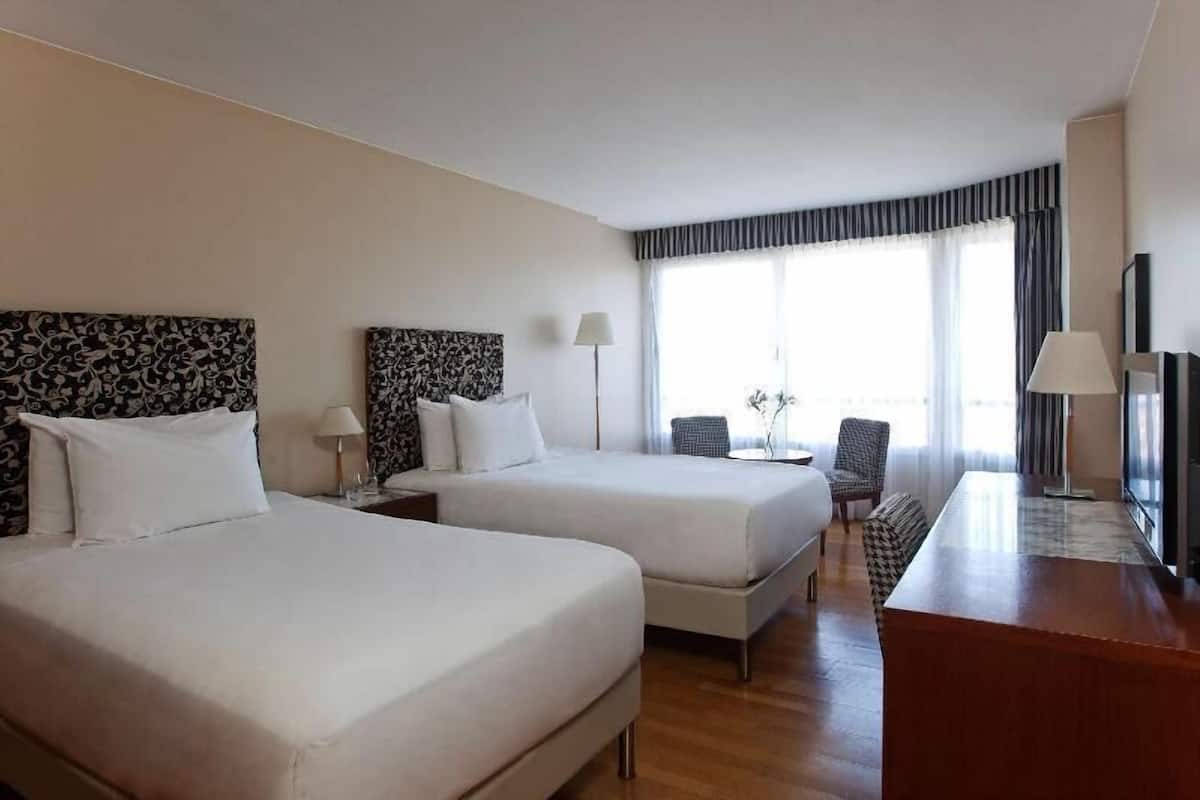 Une chambre d’hôtel avec deux lits, un bureau, une chaise et une grande fenêtre avec des rideaux.