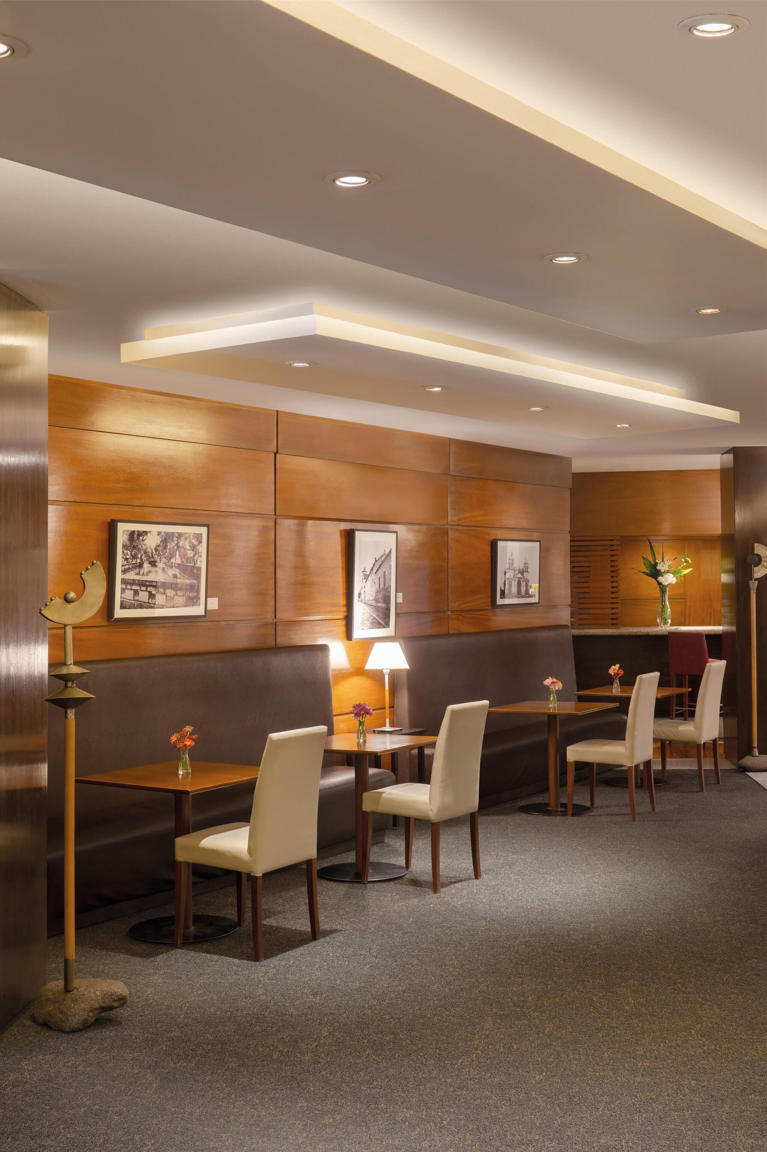 A hotel room with a dining area featuring wooden panel walls, framed pictures, a table with chairs, and a lamp.