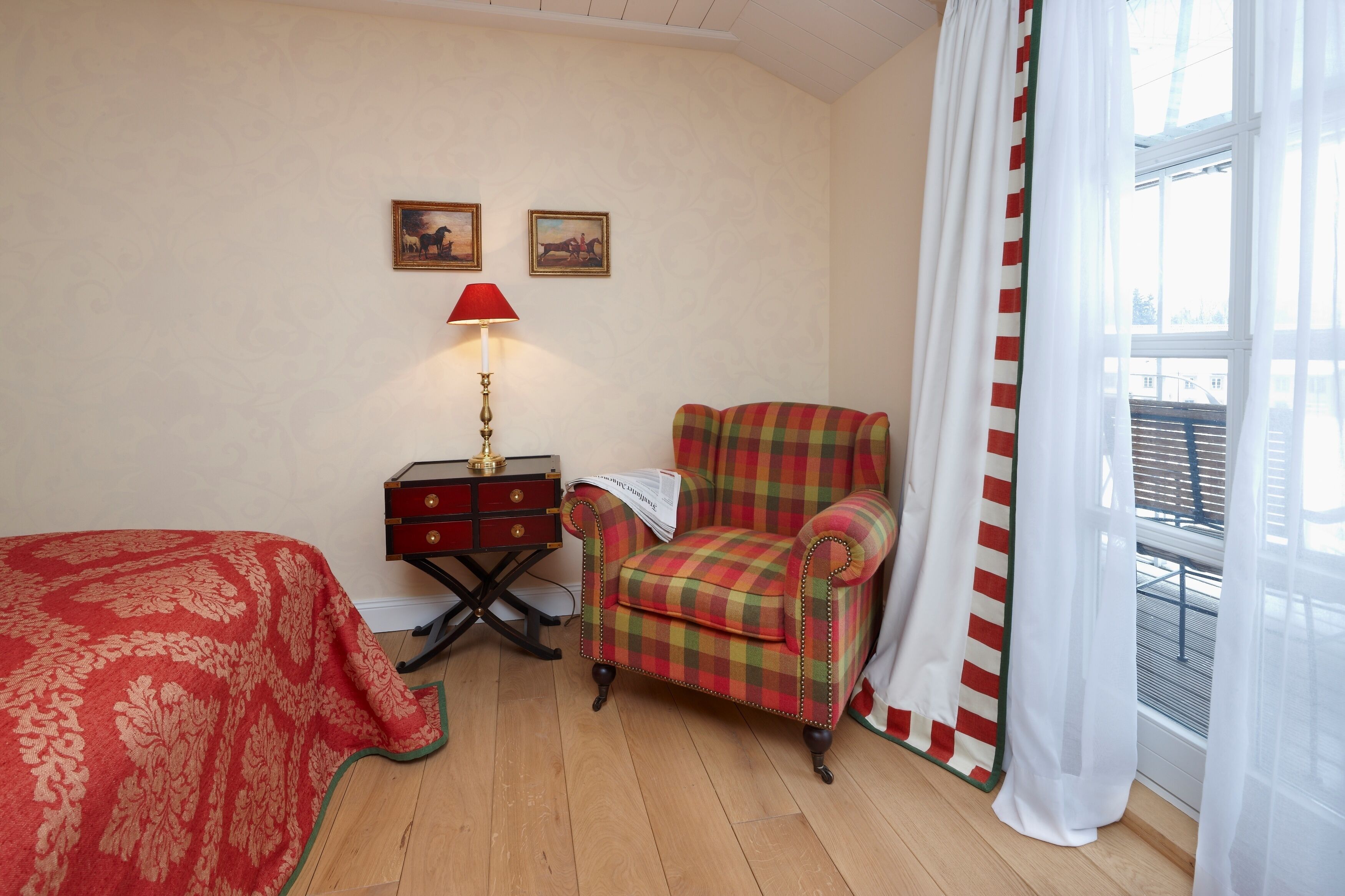 A room with a bed, a chair, a small table with a lamp, and two framed pictures on the wall.
