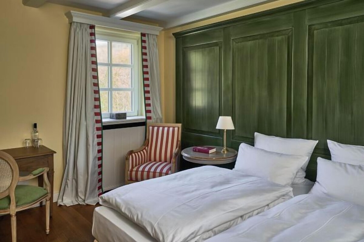 Ein Hotelzimmer mit einem Bett, einem Sessel, einem kleinen Tisch mit Lampe und einem Fenster mit Vorhängen.