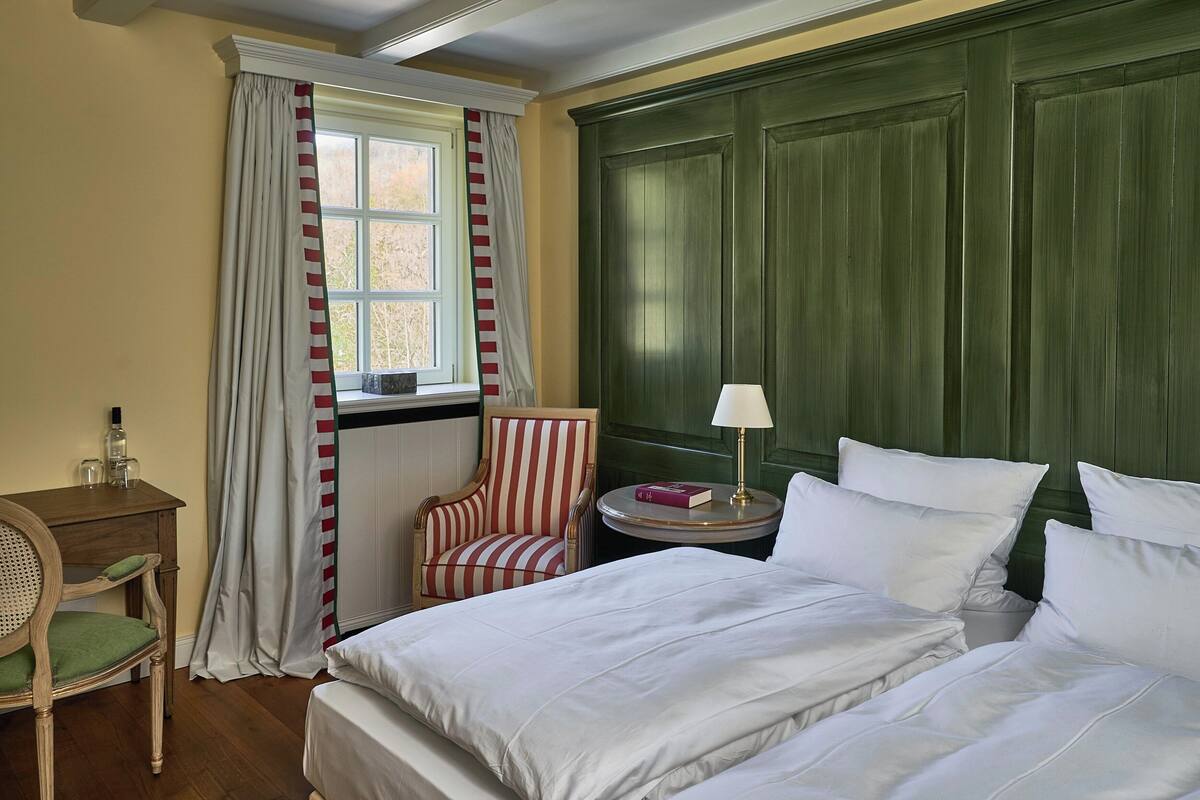 Een hotelkamer met een groot bed, een stoel, een klein tafeltje met een lamp en een raam met gordijnen.