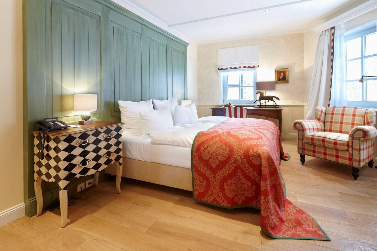 Een slaapkamer met een bed, een tafeltje met een geruit kleed, een stoel en een bureau met een lamp.