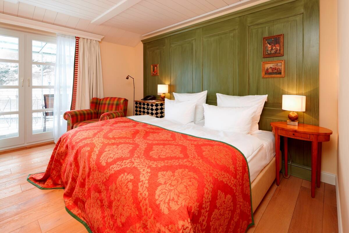 Een hotelkamer met een groot bed, een geruit fauteuil, een klein tafeltje en uitzicht op het omliggende landschap via een glazen deur.