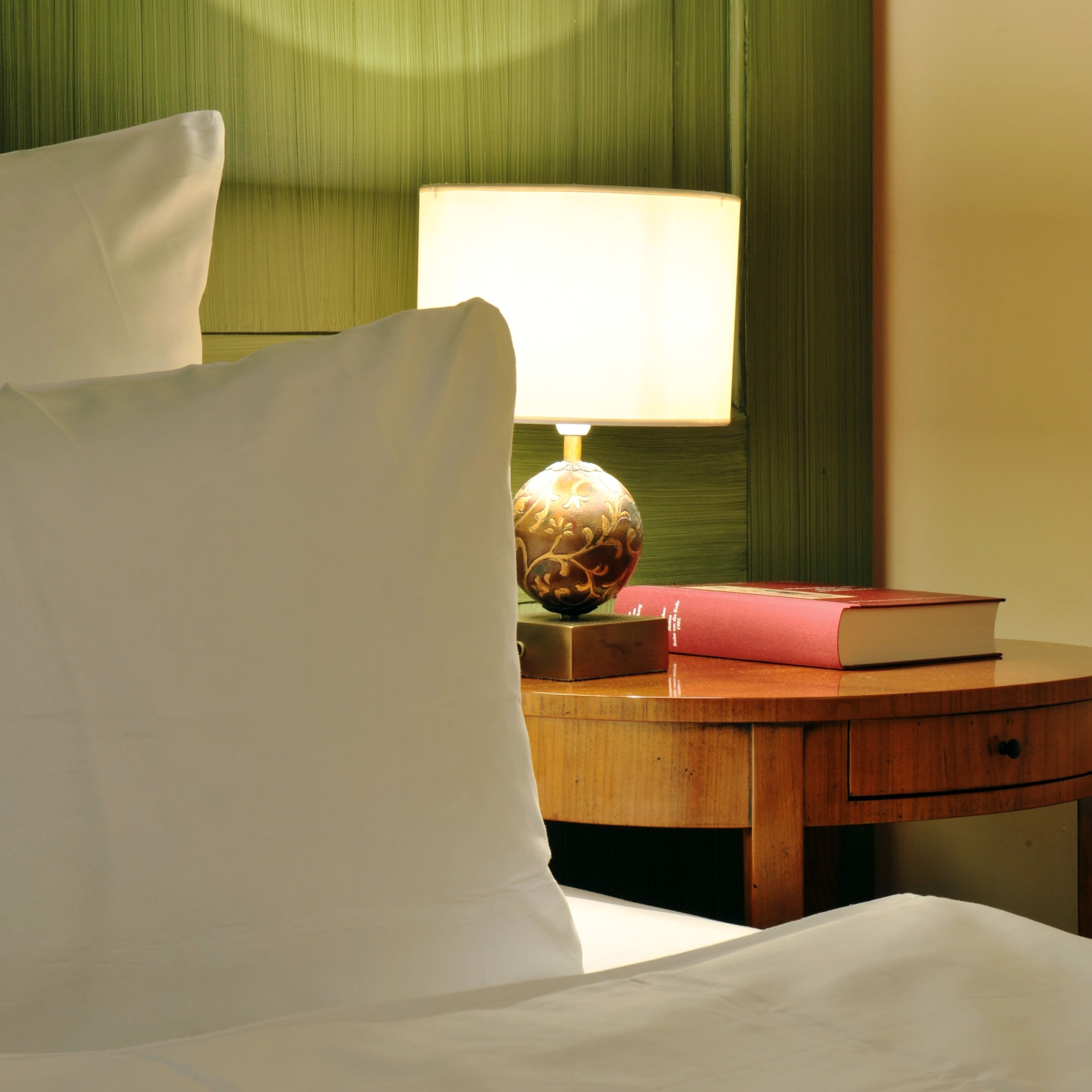 A neatly made bed with white linens, a wooden bedside table with a lamp and a book, and a green wall in the background.
