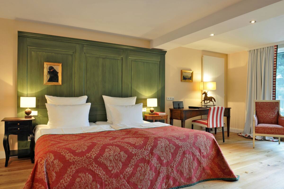 Een hotelkamer met een groot bed, twee nachtkastjes, een bureau met stoel, een lamp, en een stoel met een rood-wit gestreept kussen.