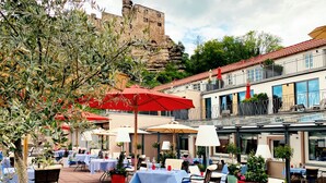 Ein Straßencafé mit Tischen und Stühlen, roten Sonnenschirmen und Blick auf ein historisches Gebäude im Hintergrund.