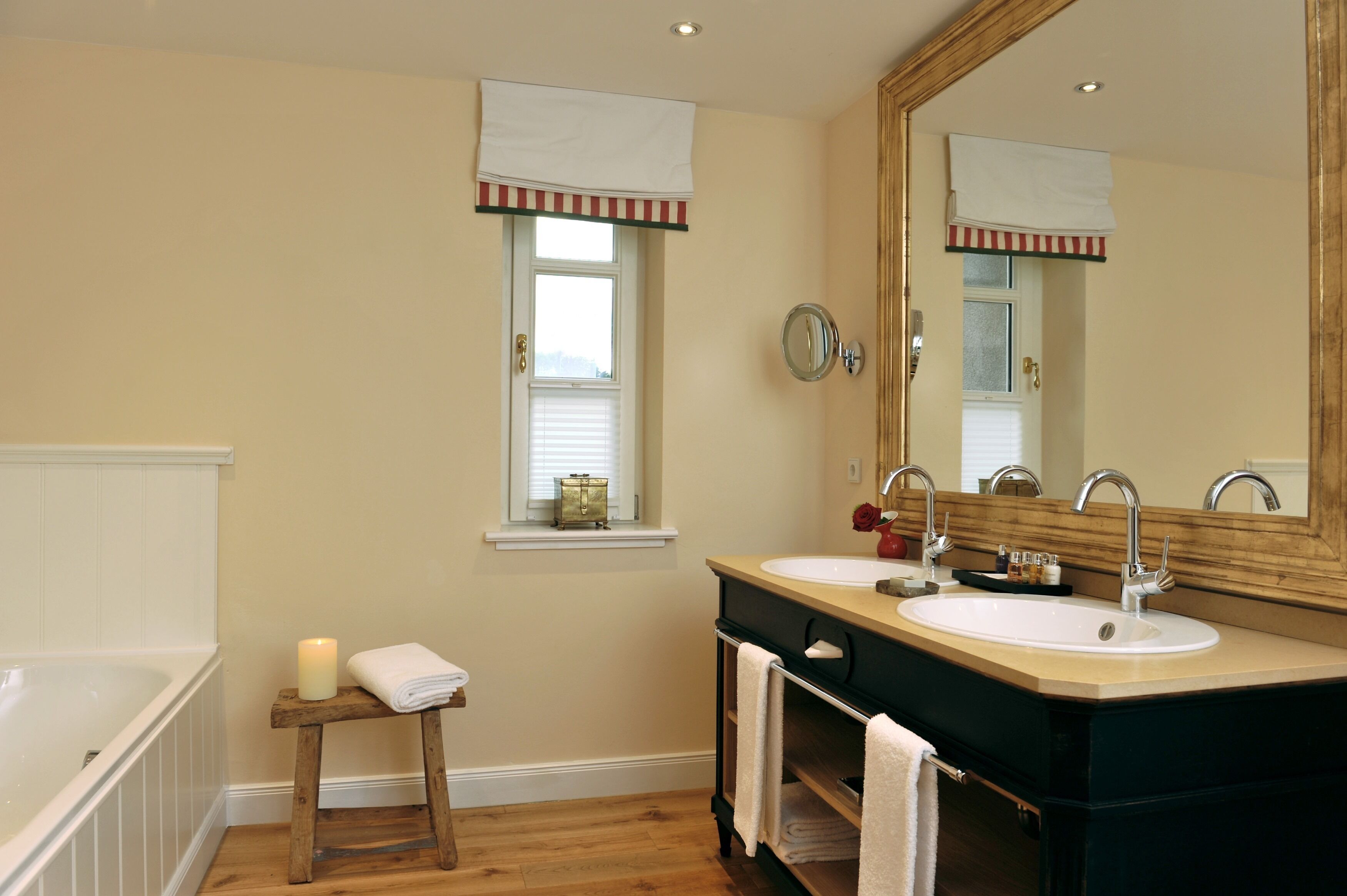 A bathroom with a double sink, a large mirror, a window with a striped valance, a small stool with a candle, and a bathtub.