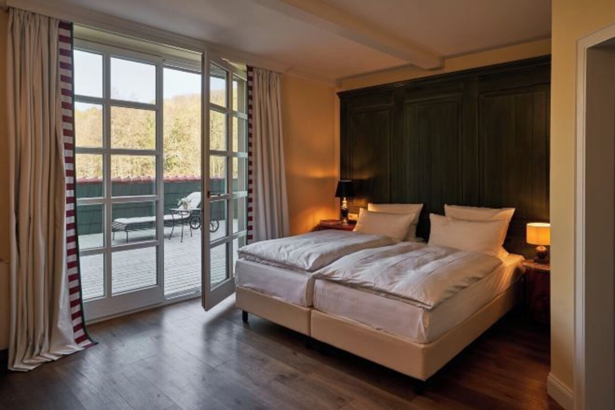 Un lit double avec du linge de lit blanc, une tête de lit en bois foncé, une table de chevet avec une lampe et une porte-fenêtre coulissante donnant sur un espace extérieur aménagé pour se détendre.