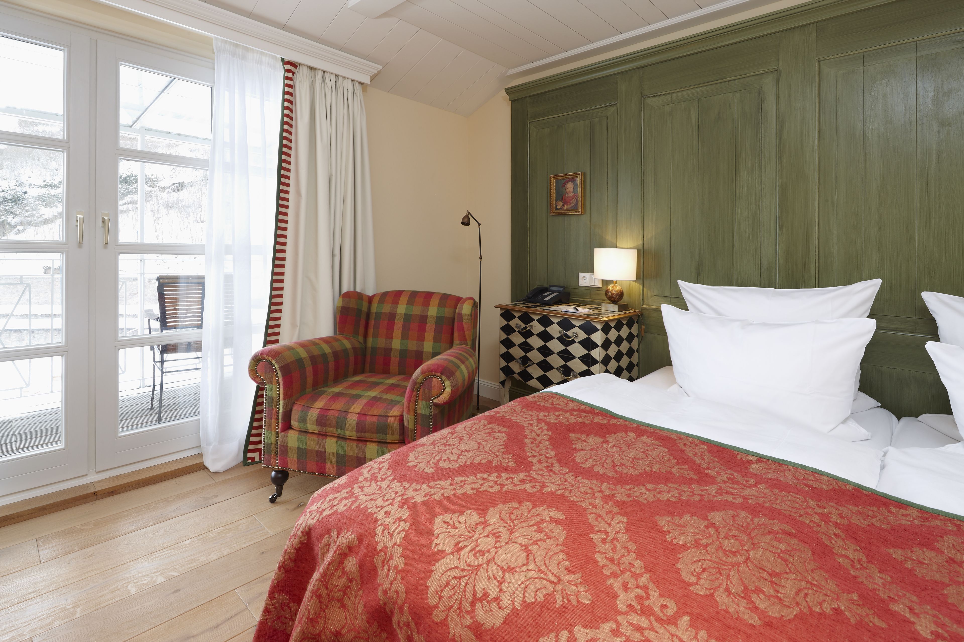 A hotel room with a bed, a plaid armchair, a checkered side table, a lamp, and a view through a glass door.