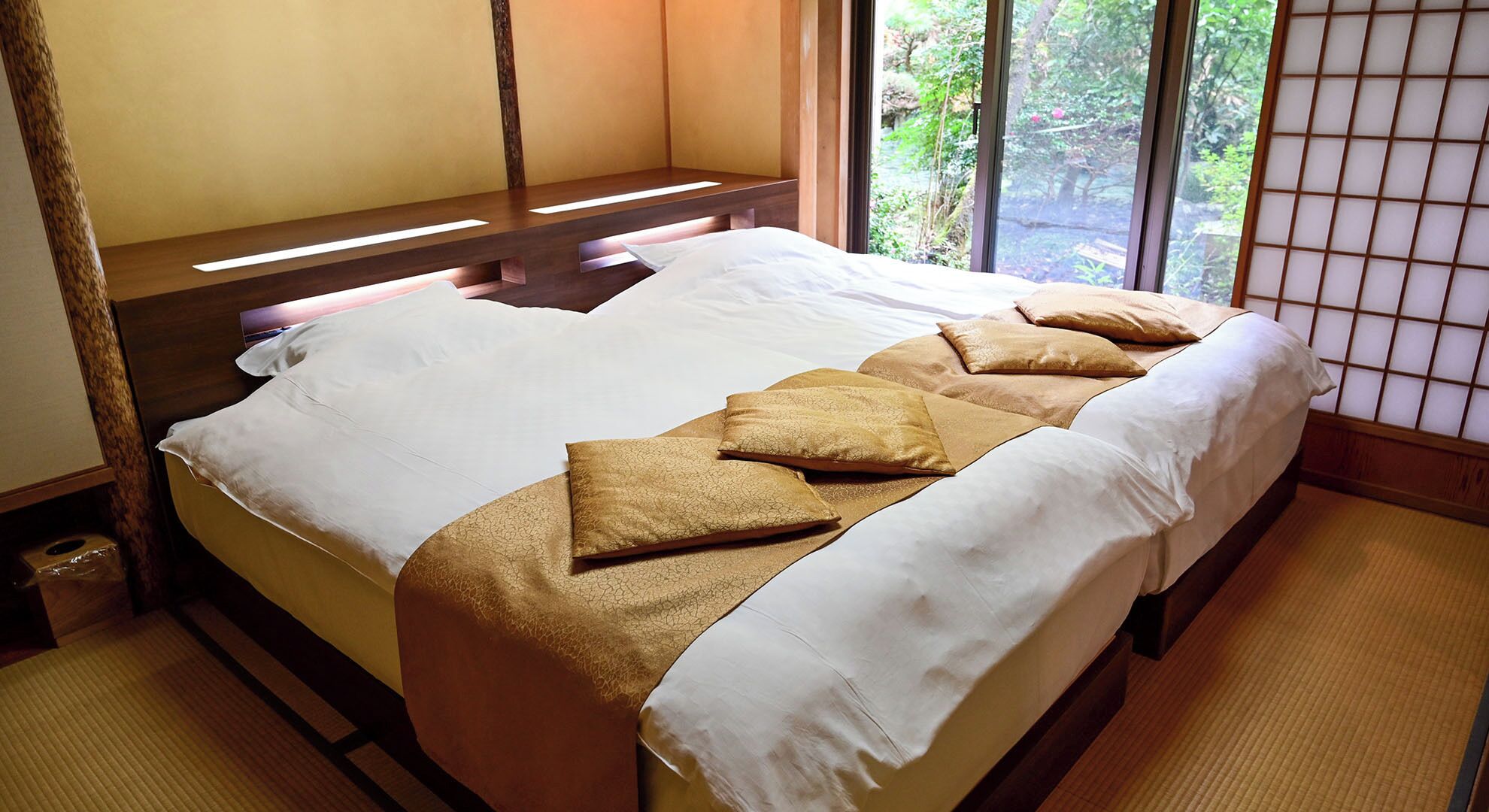 A traditional Japanese-style room with tatami flooring, a low wooden bed with white bedding, and a view of greenery through a shoji screen.