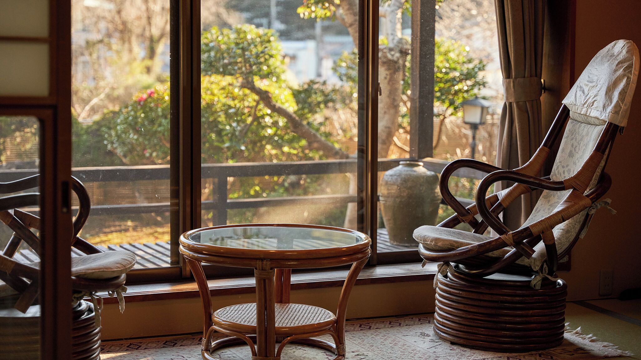 A wicker chair and a small round table by a window with a view of trees and a fence.
