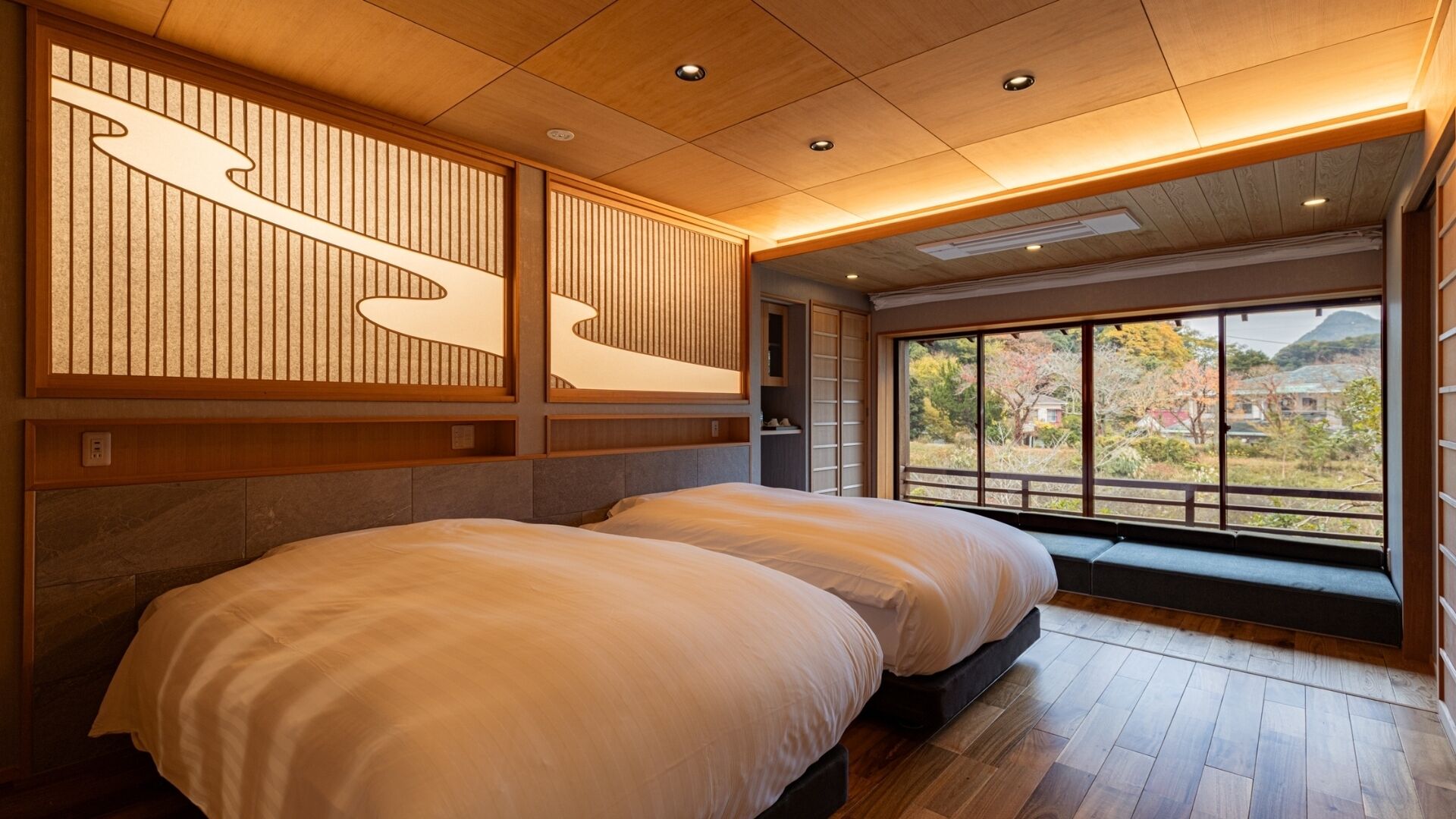 A hotel room with two beds, wooden paneling, a view of trees and buildings, and a large window.