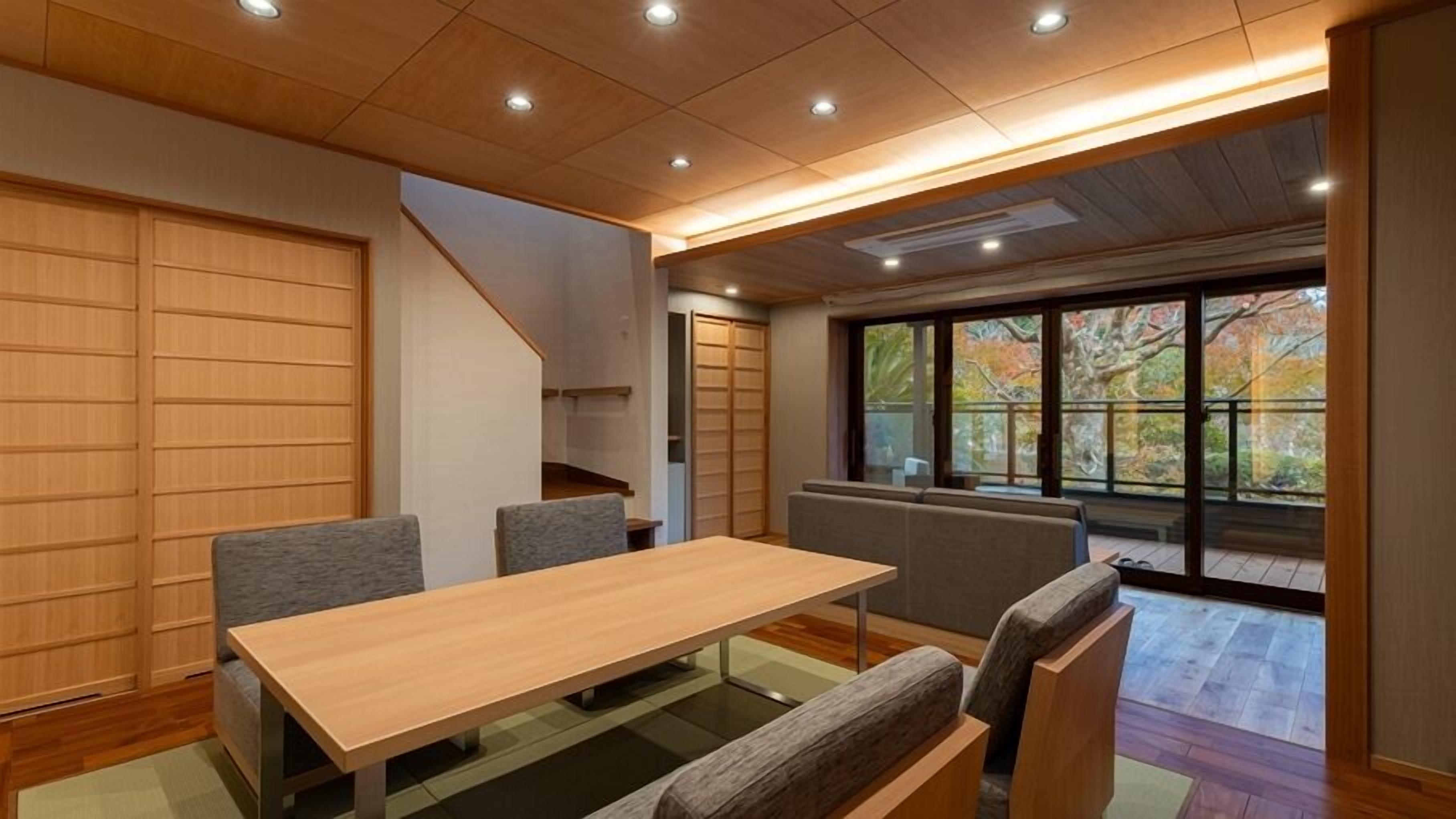 A modern interior with a wooden table and chairs, a staircase, and a large window with a view of trees.