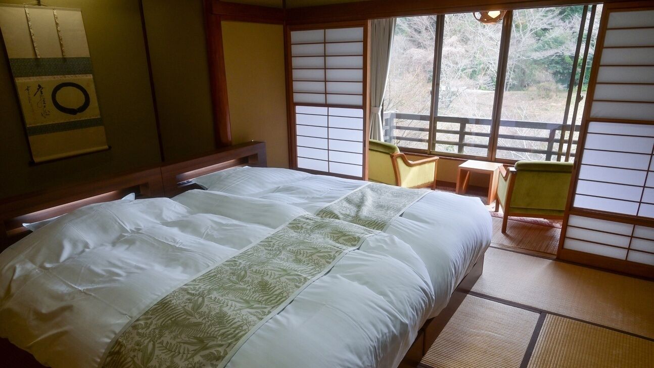 A traditional Japanese-style room with tatami flooring, a large bed with a green patterned bedspread, a small table, and sliding doors.