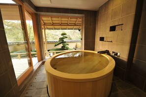 A traditional Japanese-style bathroom with a wooden bathtub, a sliding glass door leading to a garden, and a view of a bonsai tree.