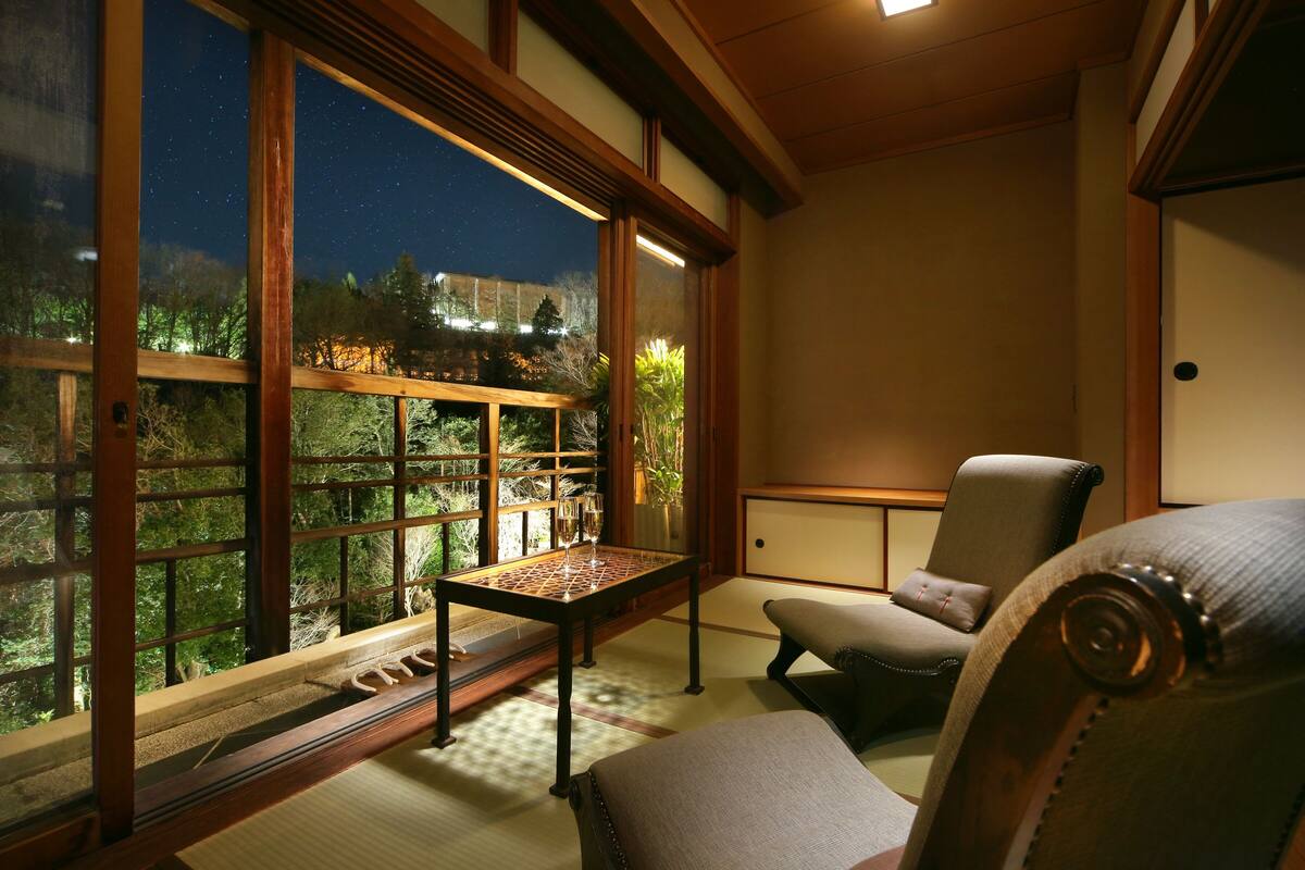 Una habitación con suelo de tatami, una mesa baja con dos vasos, un sillón acolchado y un amplio ventanal con vistas a árboles y edificios.