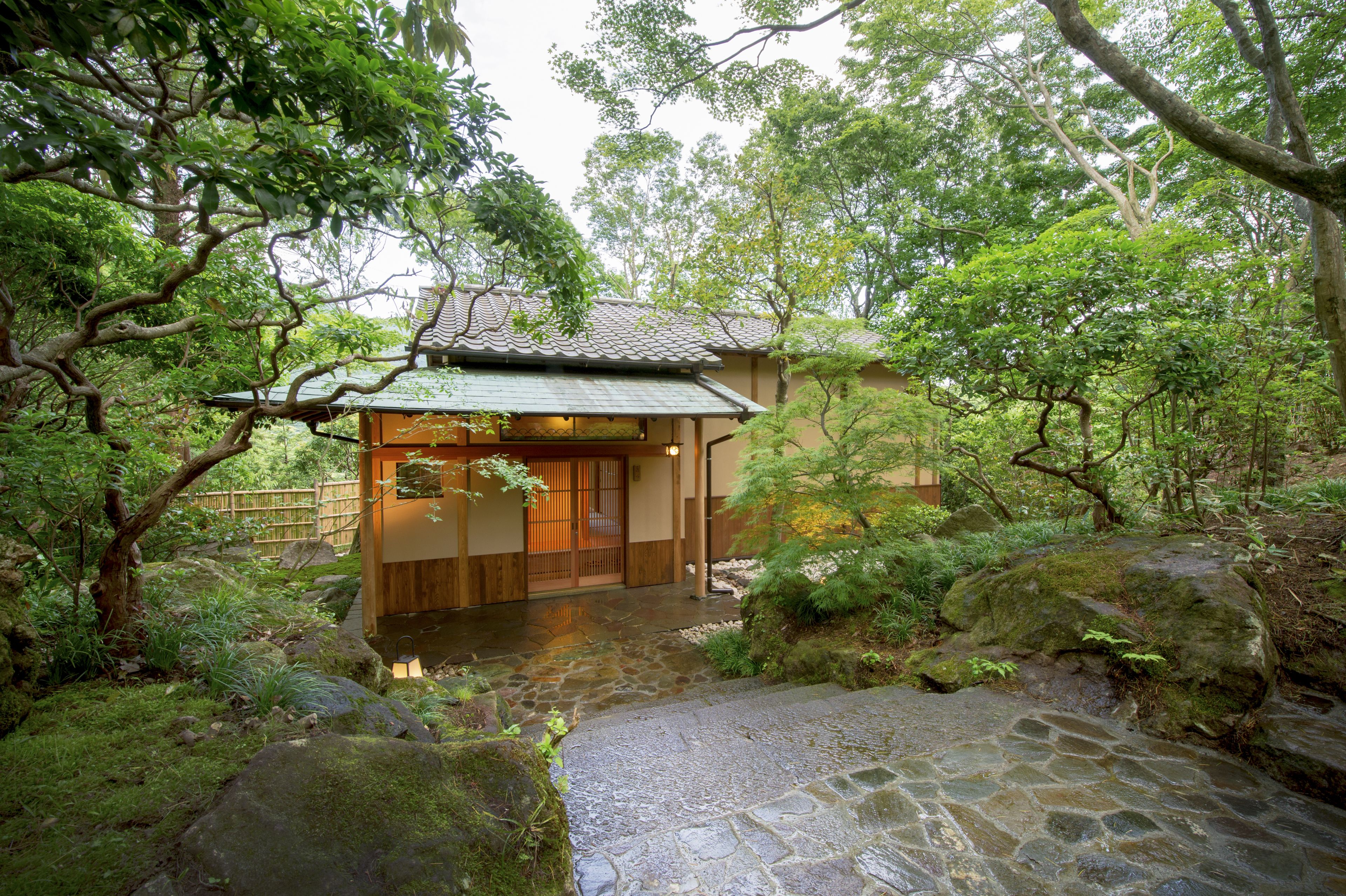 A traditional Japanese house with a stone pathway leading to the entrance, surrounded by lush greenery and a small pond.