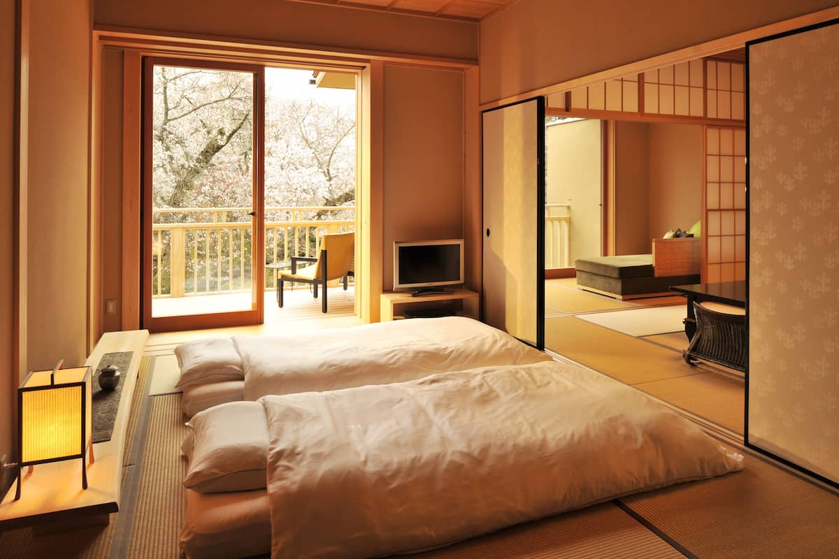 Una habitación tradicional japonesa con suelo de tatami, puertas correderas, televisor y un balcón con vistas a los cerezos en flor.