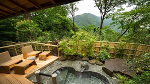 Un jacuzzi rodeado de mobiliario de madera para terraza y una piscina de piedra, ubicado en un jardín exuberante con montañas al fondo.