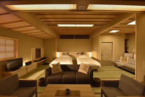A traditional Japanese-style room with tatami flooring, a low wooden ceiling, and a futon bed. There is a television on a low table and a futon sofa with cushions.