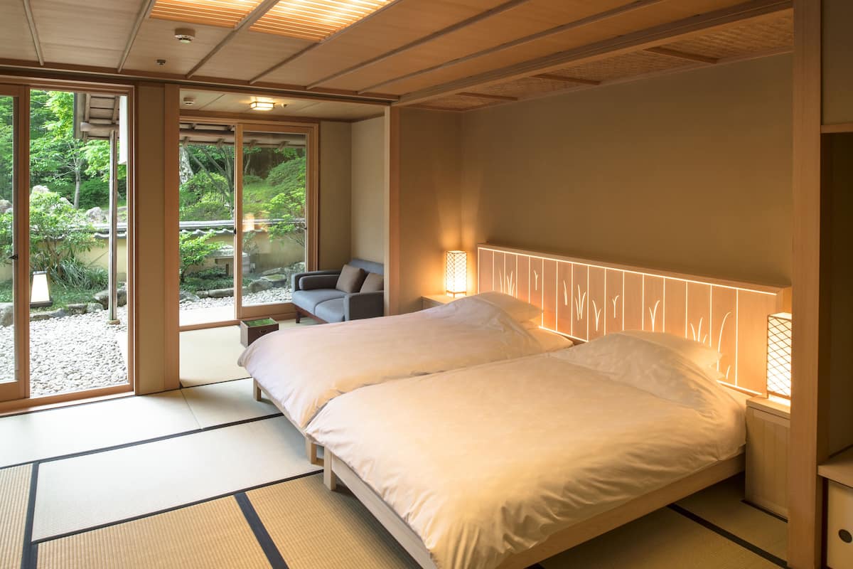 Un dormitorio de estilo japonés tradicional con suelo de tatami, dos camas, una puerta corrediza que da a un jardín y vistas de vegetación.