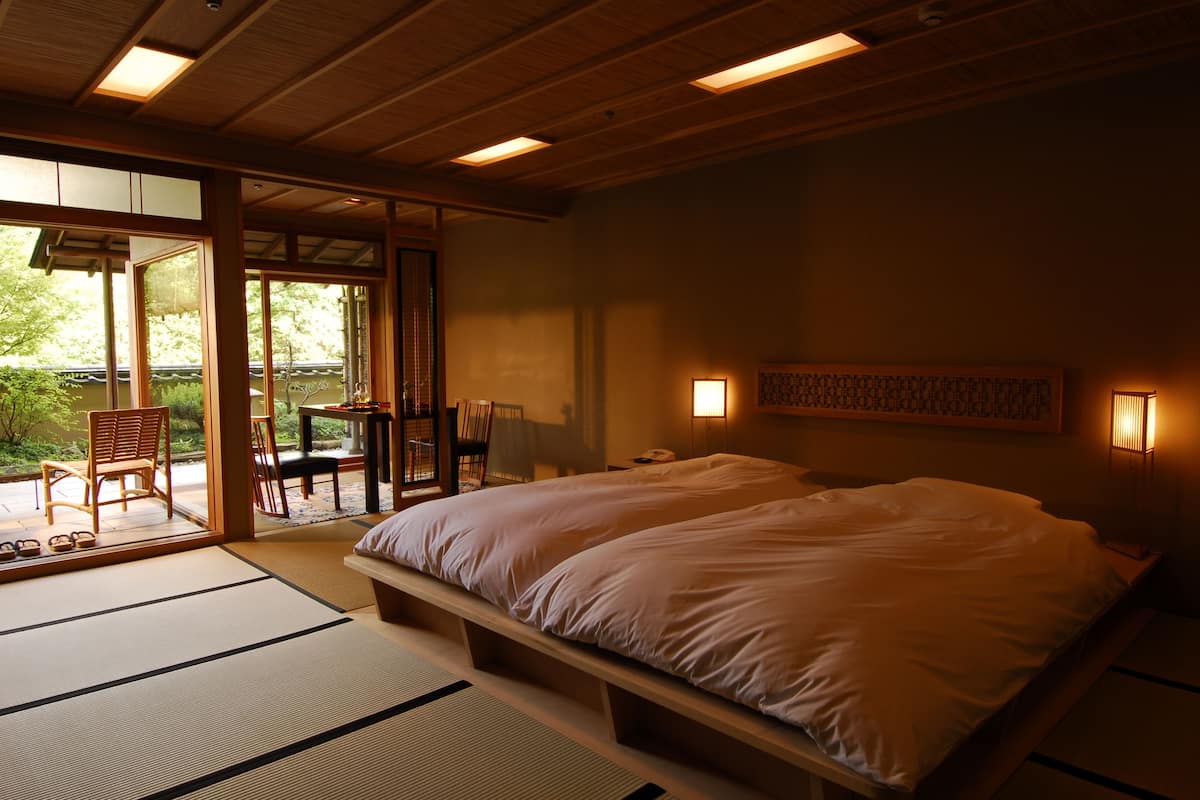 Un dormitorio tradicional japonés con suelo de tatami, una cama grande, puertas correderas que dan a un espacio exterior y vistas a la vegetación.