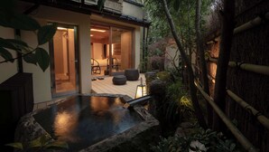 Un interior japonés tradicional con suelo de tatami, una puerta corredera que da a otra habitación y una pequeña zona de aguas termales al aire libre con rocas y plantas.