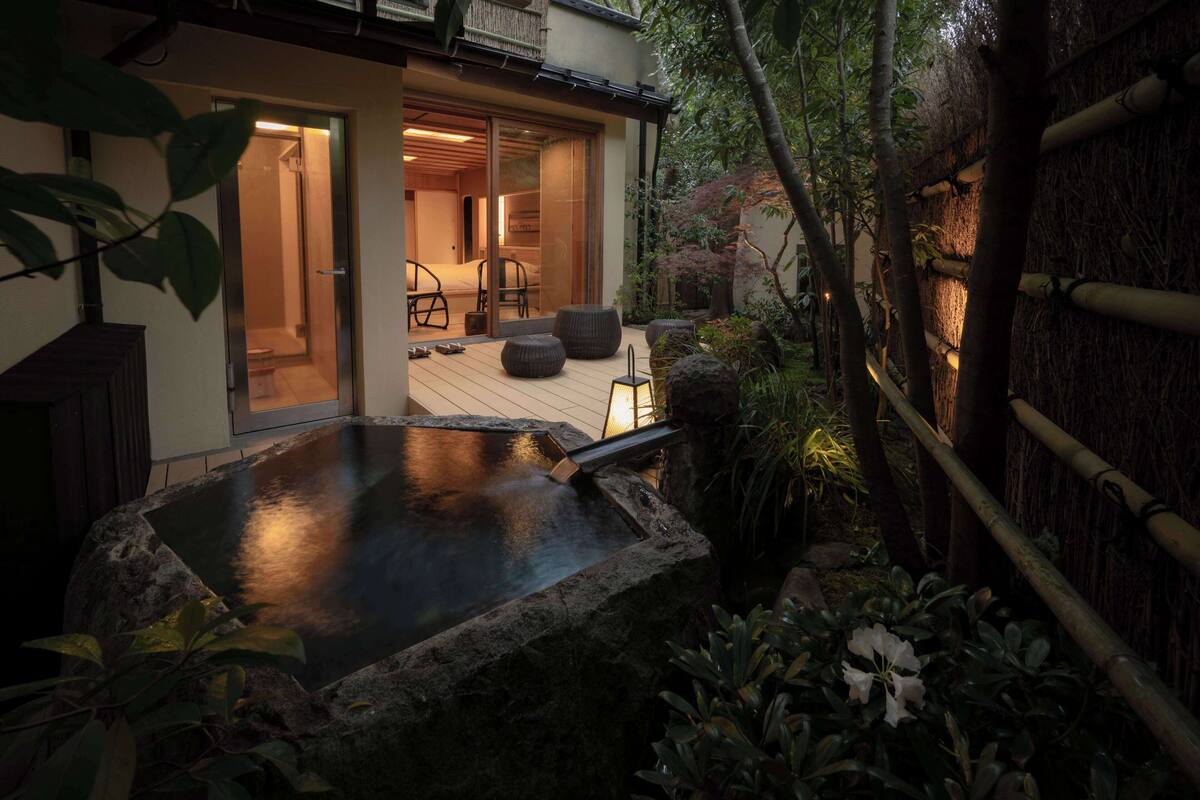 Un interior japonés tradicional con suelo de tatami, una puerta corredera que da a otra habitación y una pequeña zona de aguas termales al aire libre con rocas y plantas.