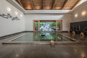A serene indoor pool area with a view of a natural waterfall, wooden beams on the ceiling, and a stone floor.