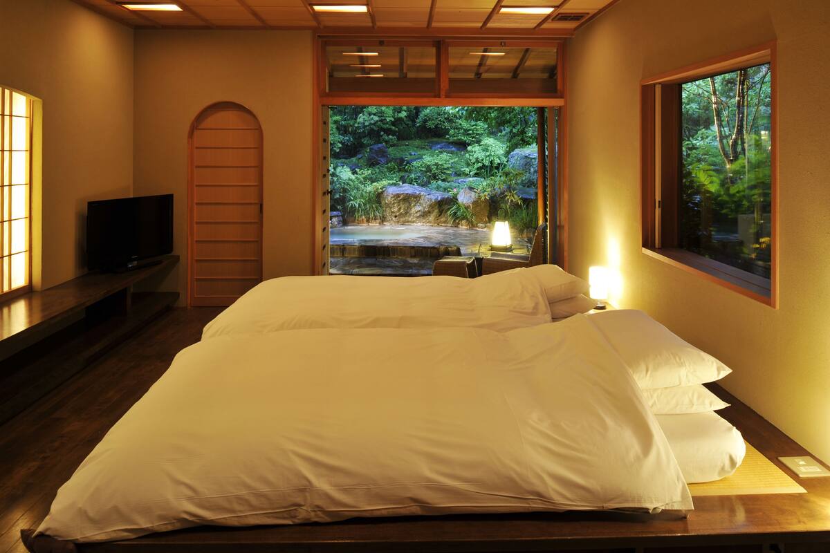 Un dormitorio con una cama grande, un televisor sobre un mueble, una ventana con vistas a un bosque y un techo de madera.
