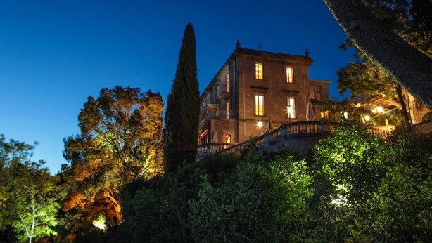 A large, historic building with multiple windows and a balcony, surrounded by dense foliage.